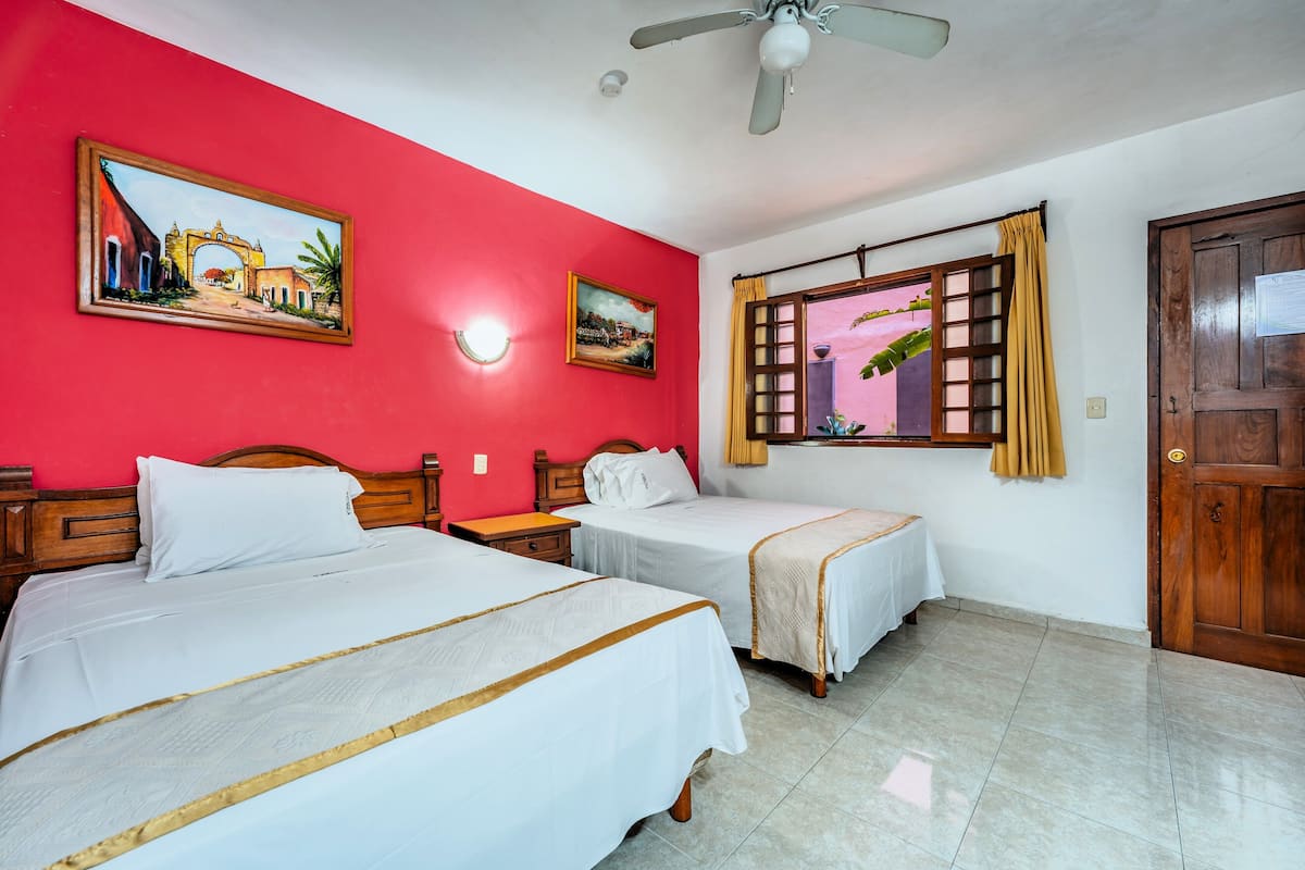A room with two beds, a ceiling fan, a window with curtains, and two paintings on the wall.