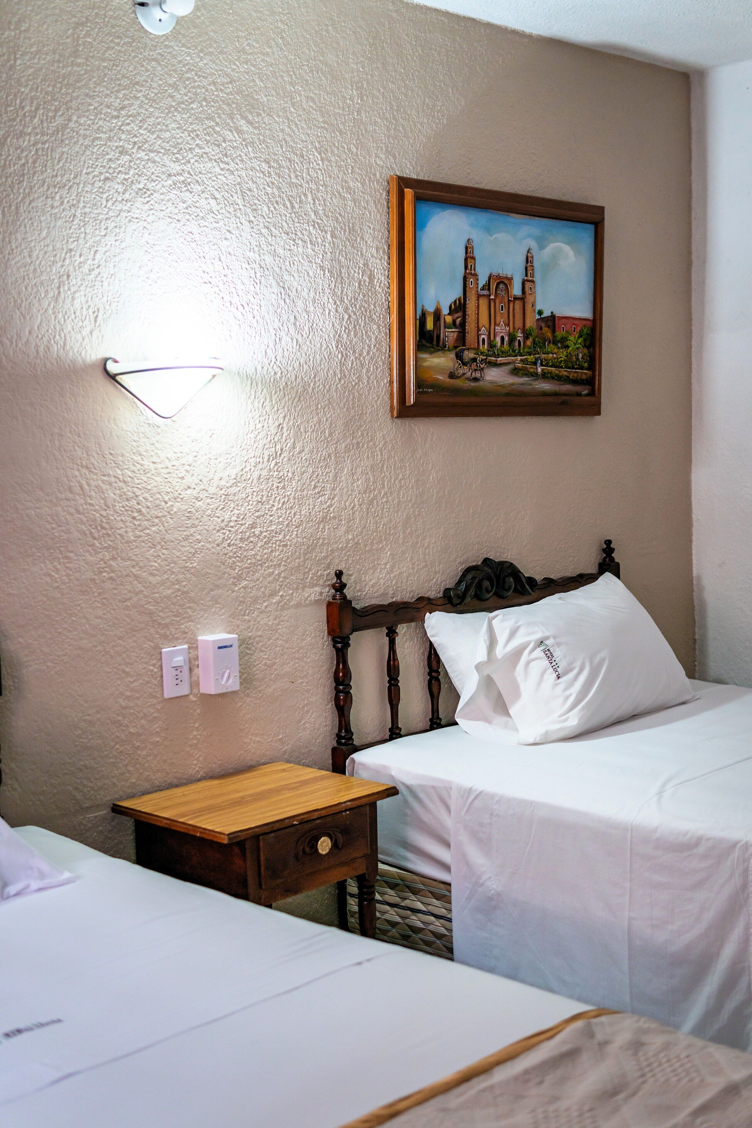 Habitación de hotel con dos camas, una mesita de noche de madera, una lámpara de pared y un cuadro de un edificio.