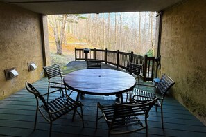 A covered outdoor patio with a round table and six chairs, surrounded by a wooden fence, overlooking a forested area.