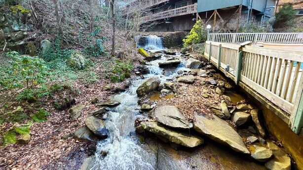 Un arroyo serpentea por un área boscosa, con un puente de madera y un edificio al fondo.