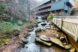 Un arroyo serpentea por un área boscosa, con un puente de madera y un edificio al fondo.