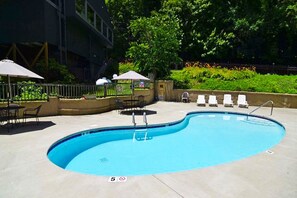 A swimming pool with lounge chairs and umbrellas, surrounded by greenery and a building.