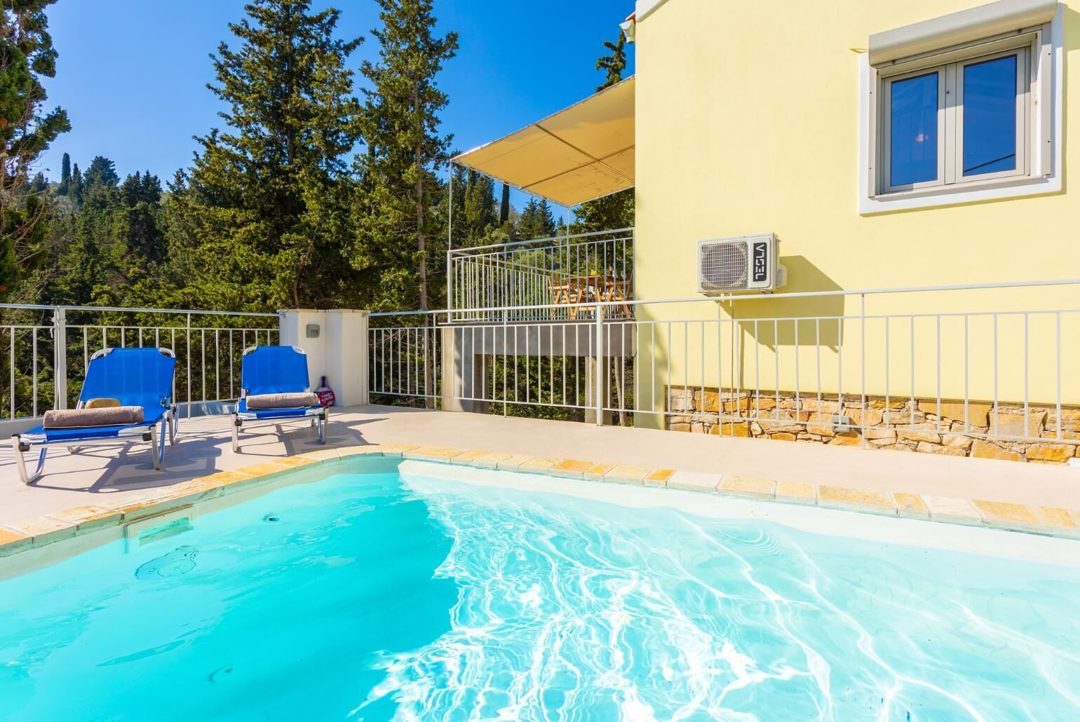Ein Schwimmbecken mit zwei blauen Liegestühlen, einer überdachten Terrasse und einem Gebäude mit Fenster und Klimaanlage.