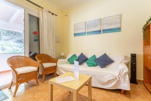 Una habitación con un sofá blanco, dos sillones de mimbre, una pequeña mesa con papeles, un ventanal con cortinas y una pared con tres cuadros.