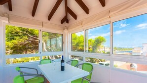 A covered patio with a table and chairs, large windows with a view of trees and buildings, and a bottle of wine on the table. - Villa Torreta - Three Bedroom Villa, Sleeps 4 (Punta Prima)