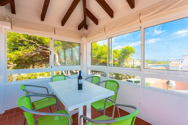Un patio techado con una mesa y sillas, ventanas amplias con vistas a árboles y edificios, y una botella de vino sobre la mesa.