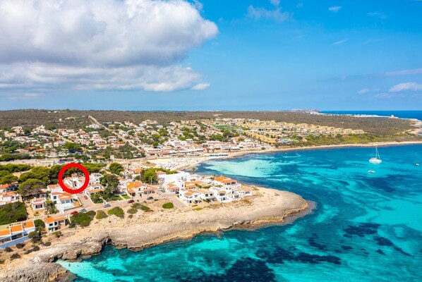 Uma cidade costeira com edifícios ao longo da orla, um mar azul e límpido, e um veleiro ao longe.