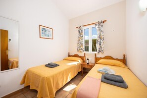 Ein Schlafzimmer mit zwei Betten, einem Fenster mit blumigen Vorhängen, einem Nachttisch mit Flasche und Glas sowie einem gerahmten Bild an der Wand.