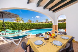 A poolside dining area with a table set for two, a pool, and lounge chairs.