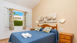 Un dormitorio con cama, dos almohadas, una mesita de noche, una lámpara de pared, un cuadro y una ventana con cortinas.