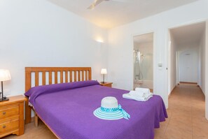 Ein Schlafzimmer mit einem hölzernen Bett, violetter Bettwäsche, einem Nachttisch mit Lampe, einem Badezimmer mit Duschkabine und einem Flur.