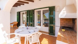 Une terrasse extérieure avec une table ronde dressée pour le repas, des volets verts et une cheminée en briques.