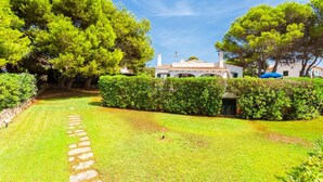Un jardin bien entretenu, avec un chemin de pierre menant à un bâtiment blanc surmonté d’un toit en tuiles.
