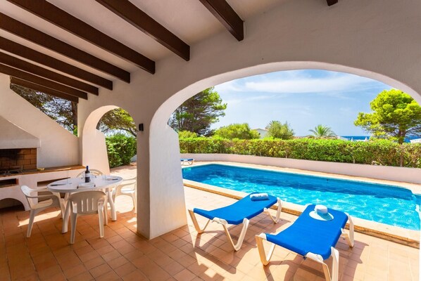 Un espace aménagé au bord de la piscine, comprenant un barbecue, une table avec des chaises et deux fauteuils de relaxation bleus.