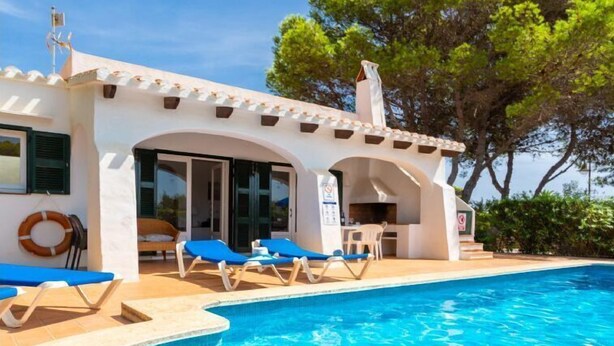 Un espace aménagé au bord d’une piscine à l’eau d’un bleu limpide, avec des chaises longues et un bâtiment à la façade blanche ornée d’arcades.