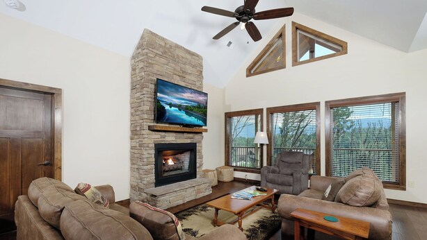 A living room with a stone fireplace, a ceiling fan, and large windows. - Relax and Soak with a Mountain View! - Bear Watch 1011 (Gatlinburg)