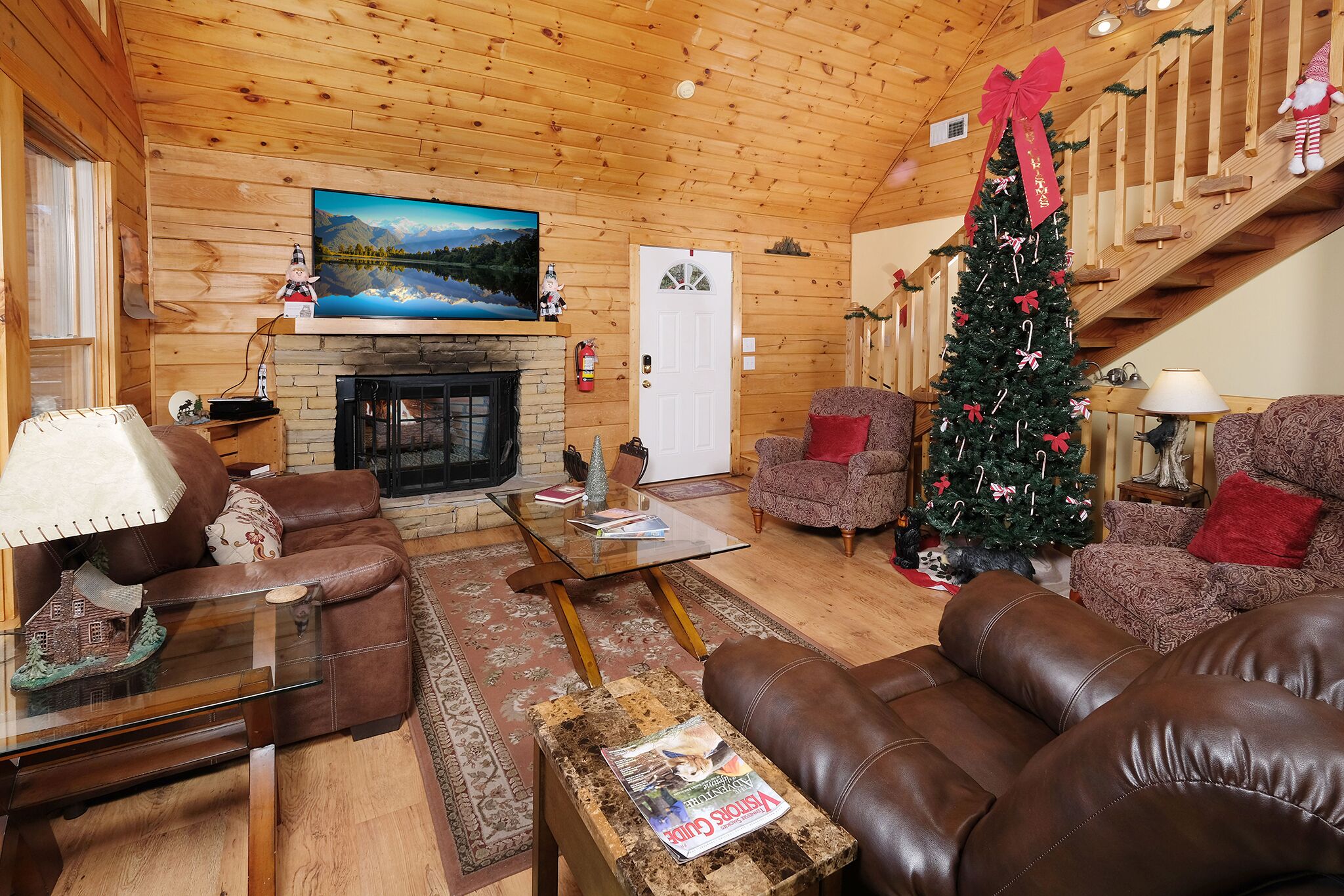 A cozy log cabin interior with a fireplace, leather sofas, a Christmas tree, and a scenic painting on the wall.