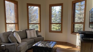 Una sala de estar con un sofá gris, un reposapiés negro y una mesa de centro de madera. Hay tres ventanas con marcos y persianas de madera, y un televisor sobre un mueble de madera.