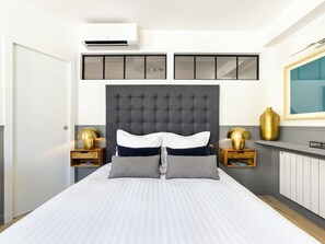 A modern bedroom with a large bed, two bedside tables with lamps, a wall-mounted air conditioner, and a large mirror.