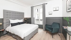 Une chambre moderne avec une tête de lit capitonnée grise, une couvre-lit blanche, un fauteuil vert foncé et une vue sur un balcon à travers une fenêtre ornée de rideaux gris.