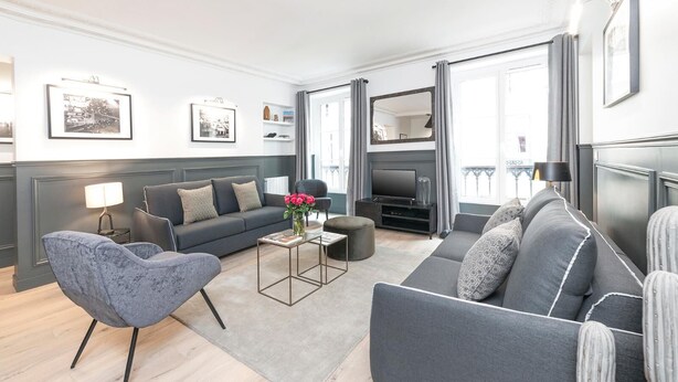 Un salon moderne avec un canapé gris, un fauteuil bleu et une table basse. On y trouve un téléviseur à écran plat posé sur un meuble, une lampe et des tableaux encadrés accrochés au mur.