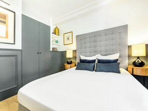 Una habitación de hotel con una cama grande, cabecera gris, dos mesitas de noche con lámparas y un armario.