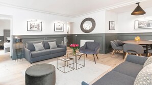 Un salon moderne avec un canapé gris, un pouf rond et une table basse. Des tableaux encadrés ornent le mur, et une plante, un cactus, est également présente.