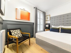 A hotel room with a bed, a chair, a radiator, a window, and a wall-mounted artwork. - LivinParis | Luxury AC 3 Bedrooms Le Louvre III (Paris)
