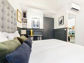 A hotel room with a large bed, a bedside table, a lamp, a bookshelf, and a door leading to another room. - LivinParis | Luxury AC 3 Bedrooms Le Louvre III (Paris)