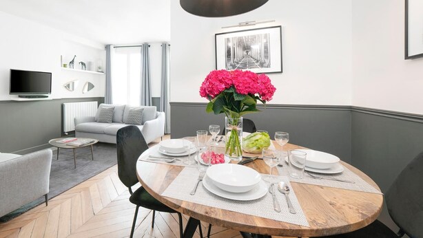 Un espace repas moderne avec une table ronde en bois, dressée pour quatre personnes et entourée de chaises. Un grand luminaire suspendu noir est fixé au plafond, et une photographie en noir et blanc, encadrée, orne le mur.