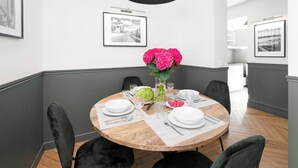 Un coin repas avec une table ronde en bois, des chaises noires et un luminaire suspendu noir. Les murs sont ornés de tableaux encadrés et un vase rempli de fleurs roses est posé sur la table.