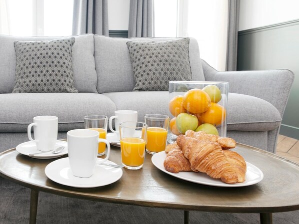 A breakfast setup with croissants, orange juice, and coffee on a table in a modern living room with a grey sofa and patterned cushions. - LivinParis | Luxury 2 Bedrooms Opera I (Paris)
