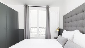 Une chambre d’hôtel moderne avec un grand lit, des rideaux gris, une tête de lit grise et une vue sur un balcon doté d’une balustrade.