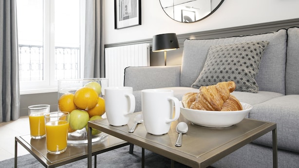 Een moderne woonkamer met een grijze bank, een ronde spiegel en een ontbijtje op een tafel, met daaronder een schaal croissants, een koffiepot en glazen sinaasappelsap.