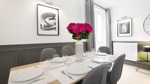 Een eetruimte met een lange tafel voor zes personen, ingelijste foto's aan de wand en een vaas met roze bloemen.