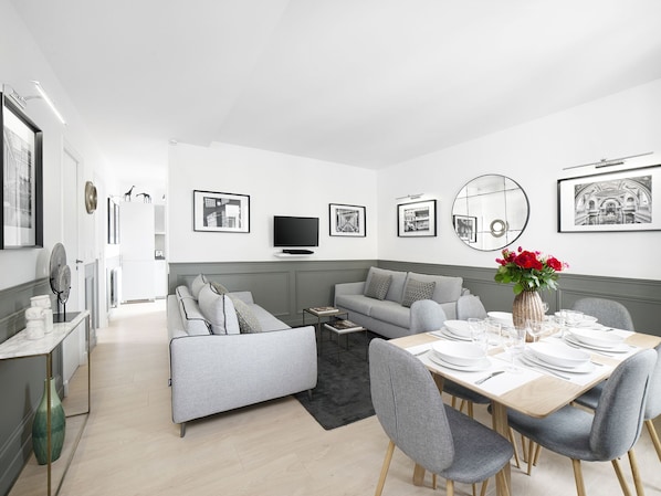 Una sala moderna con un sofá gris, una mesa de comedor para cuatro personas y un televisor de pantalla plana montado en la pared.