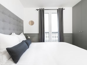 Un dormitorio moderno con una cama grande, cabecera gris, ropa de cama blanca y cortinas grises.