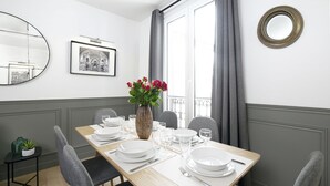 Ett matrum med ett träbord dukat för fyra personer, grå stoppade stolar, en vas med blommor, en inramad tavla på väggen och ett fönster med gardiner.