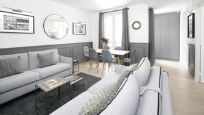 Ett modernt vardagsrum med en grå soffa, ett soffbord och en matplats med ett bord och stolar. På väggen hänger inramade tavlor och det finns även en spegel.