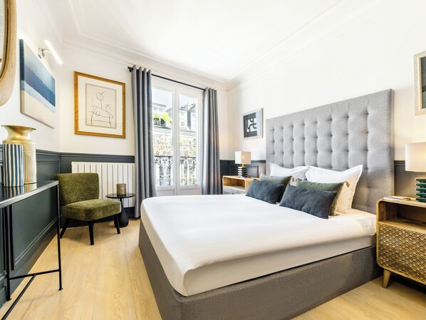 A hotel room with a large bed, a chair, a bedside table, a lamp, and a view of the outside.