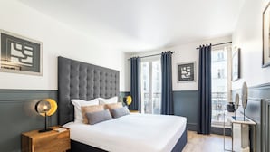 Quarto com cama grande, mesa de cabeceira com abajur, radiador e janela com cortinas.