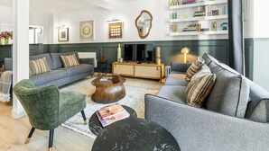 Uma sala de estar moderna com um sofá cinzento, uma poltrona verde, uma mesa de centro preta e um aparador de madeira.