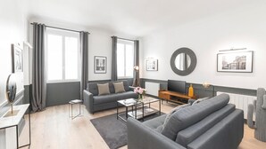Ein modernes Wohnzimmer mit grauem Sofa, einem Couchtisch und einem Fernseher. Zwei Fenster mit Vorhängen, ein runder Spiegel und gerahmte Bilder schmücken die Wand.