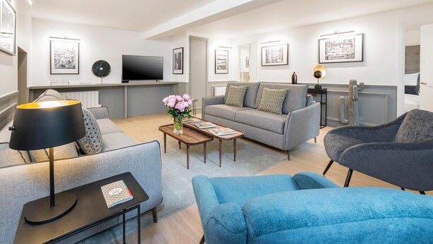 Ett modernt vardagsrum med en grå soffa, en blå fåtölj och ett soffbord med en vas med blommor. En platt-TV är monterad på väggen och en lampa står på ett sidobord.
