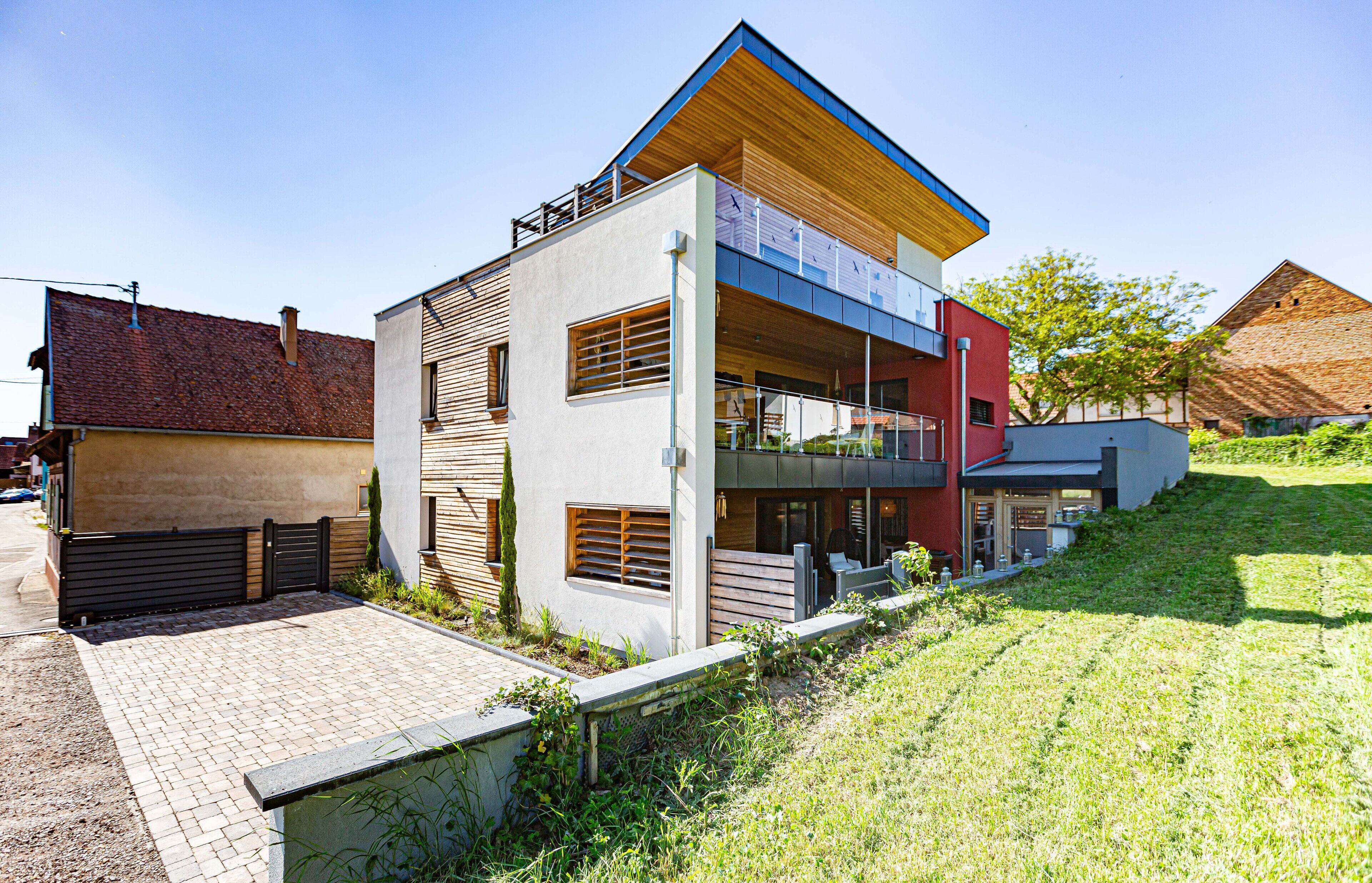 A modern two-story house with a flat roof, a balcony on the upper floor, and a paved driveway leading to the entrance.