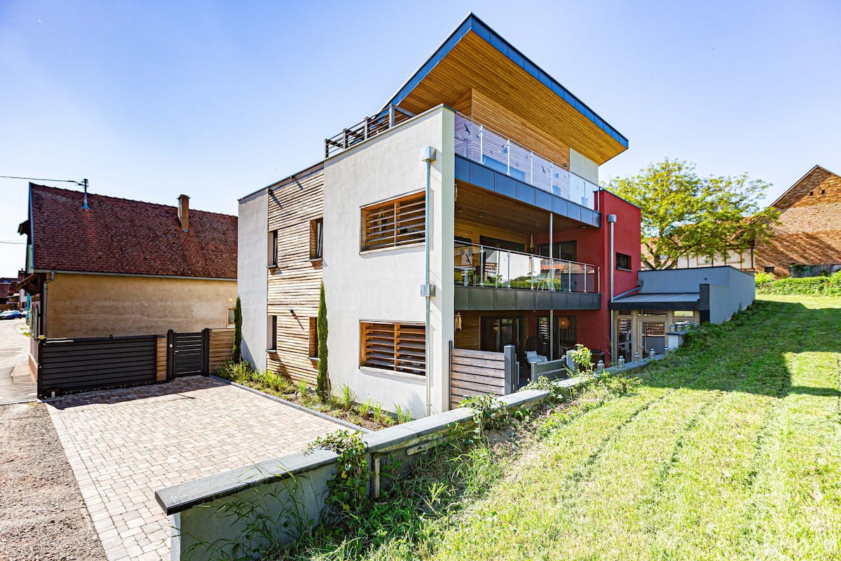 A modern two-story house with a flat roof, a balcony on the upper floor, and a paved driveway leading to the entrance.