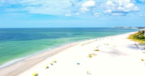 A beach with white sand, clear turquoise water, and a few umbrellas. - 🐚BBR #10, Steps to the Beach, HTD Pool, Lrg Rm w/Living Rm, Kitchen & 4 Beds! (Treasure Island)