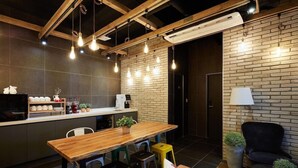 A modern kitchen with a wooden table, bar stools, and a black chair. There is a coffee machine and a potted plant on the counter. - Hostel Cleo Seoul Hongdae (Seoul)