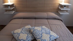 Een bed met een beige sprei en twee gestreepte kussens, met aan weerszijden nachtkastjes met lampen en een kleine plant.