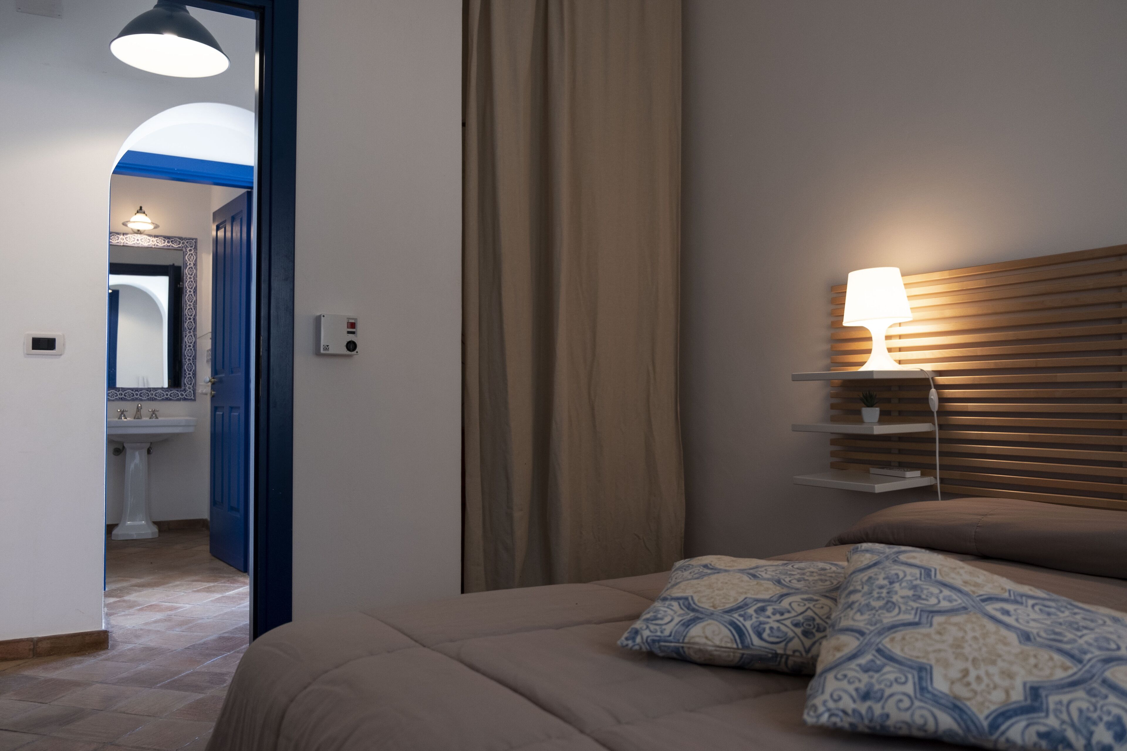 Una camera da letto con un letto, cuscini, un comodino con una lampada, una mensola a parete e una finestra con tende.